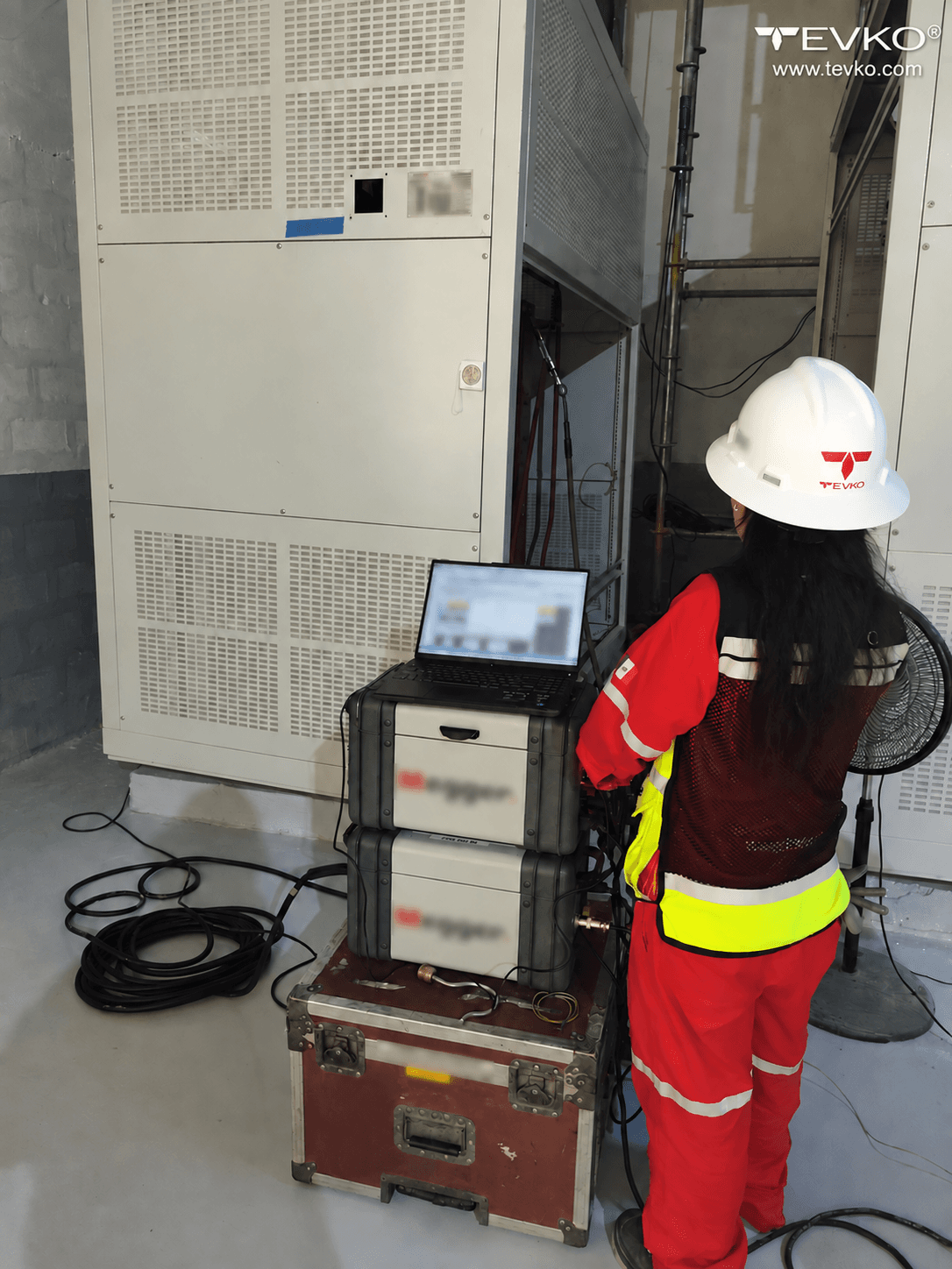 TEVKO personnel facing a transformer cabinet with a laptop and electrical test instruments.