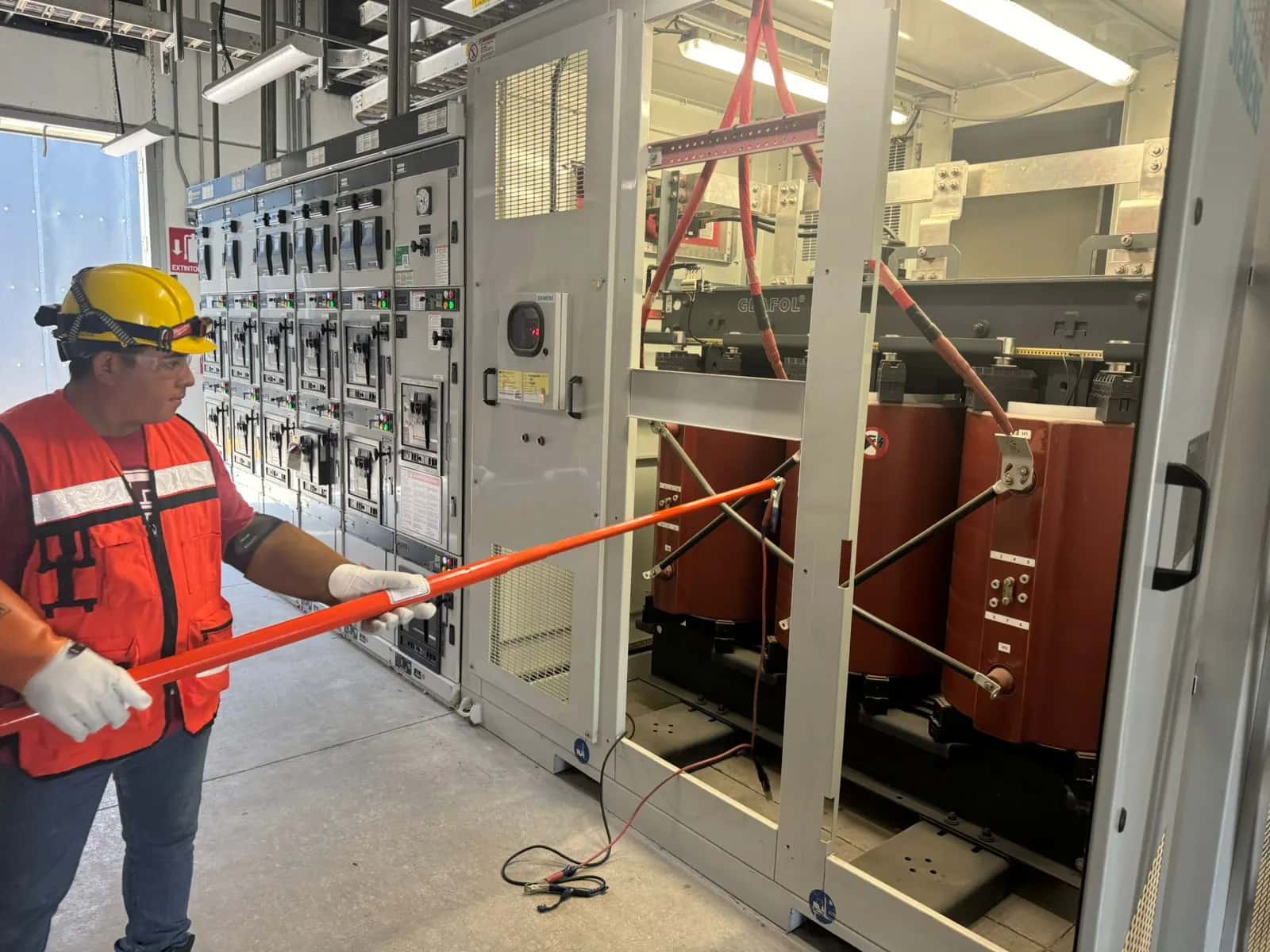 Técnico con guantes dieléctricos operando una pértiga aislante frente a un tablero de celdas de media tensión, con un transformador seco al fondo