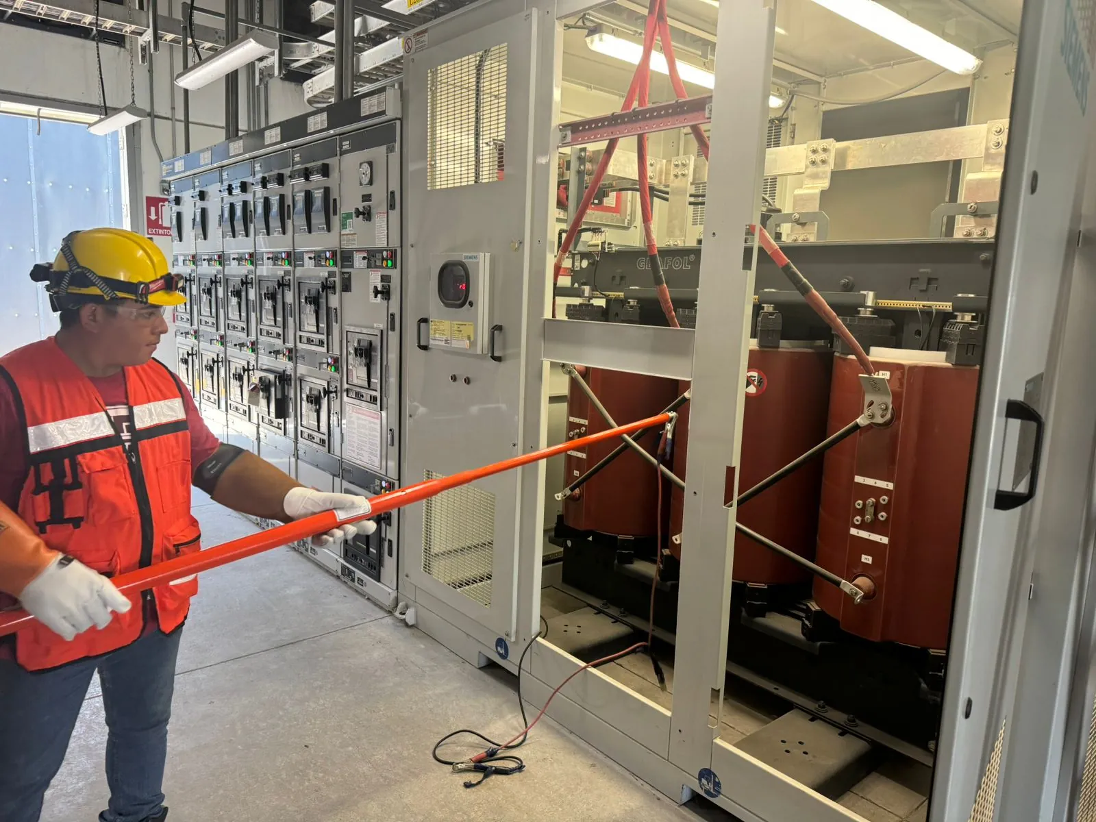 Técnico con guantes dieléctricos operando una pértiga aislante frente a un tablero de celdas de media tensión, con un transformador seco al fondo