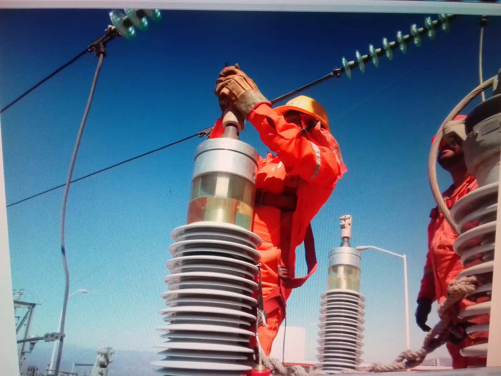 Técnico en overol naranja conectando el cable de prueba en la parte alta de una boquilla, en el patio de una subestación