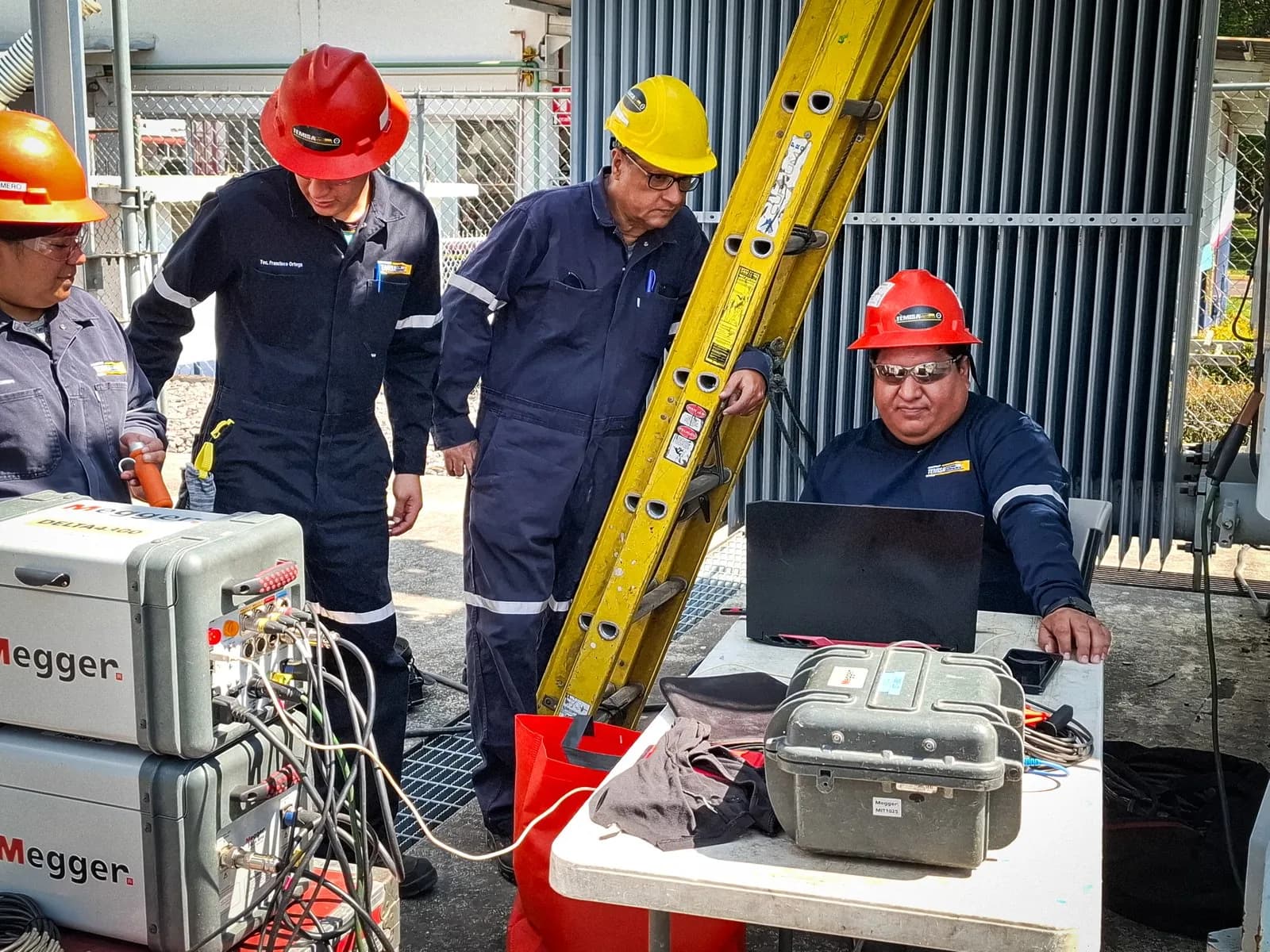 Cuadrilla de cuatro técnicos alrededor de dos instrumentos Megger y una computadora portátil, en el patio de una subestación