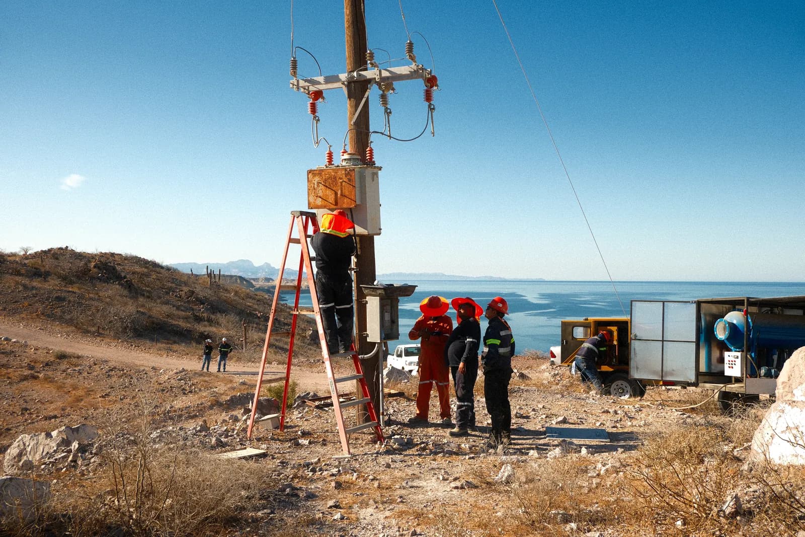 Cuadrilla con escalera de tijera trabajando en un transformador montado en poste, en terreno abierto junto a la costa