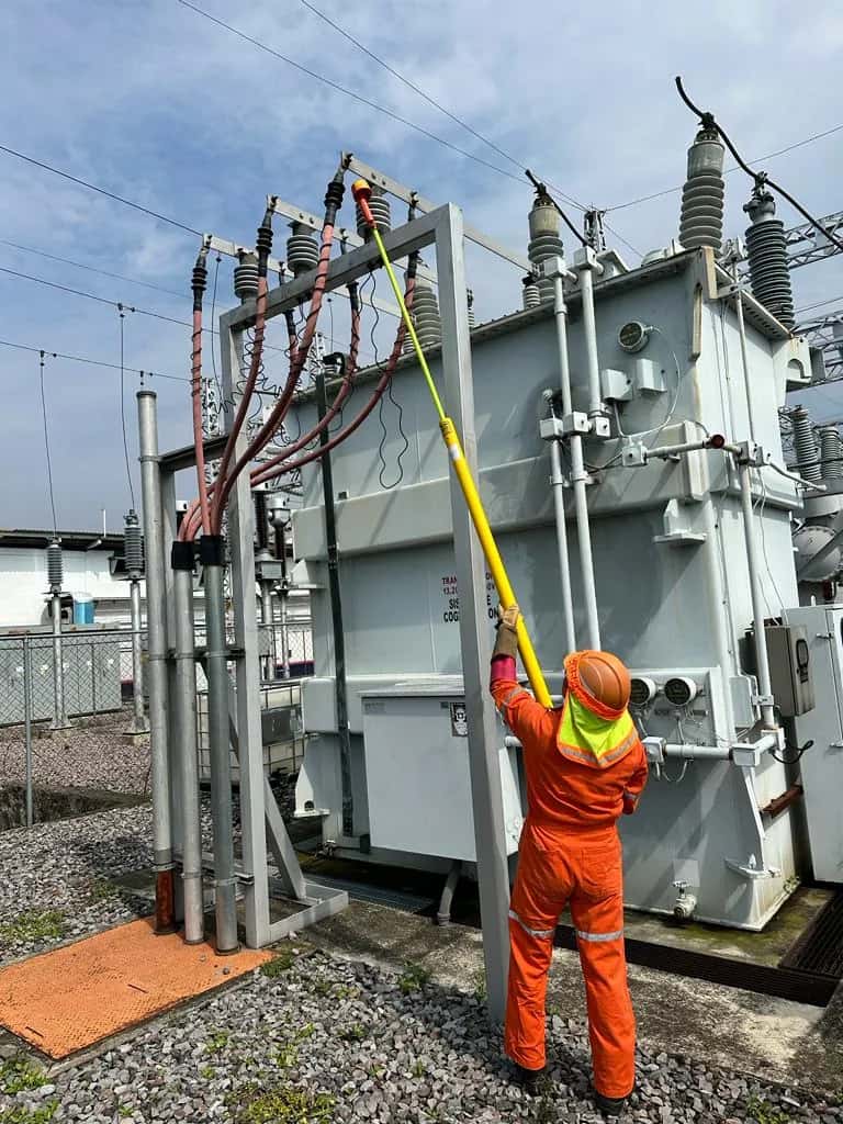 Técnico en overol naranja maniobrando con una pértiga sobre un transformador de potencia en el patio de una subestación