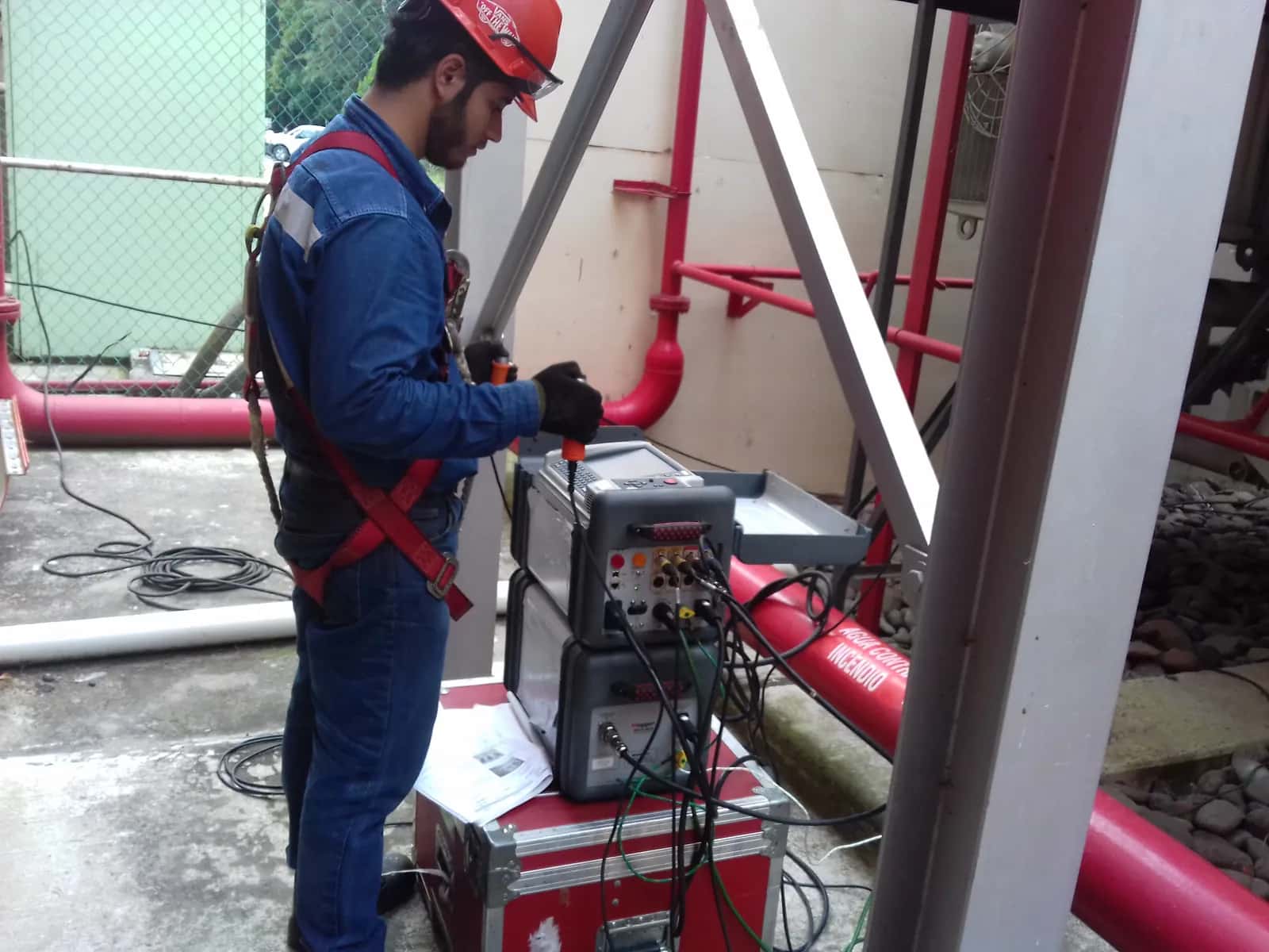Técnico con arnés operando el equipo de prueba de factor de potencia sobre una plataforma, junto al estuche y la computadora
