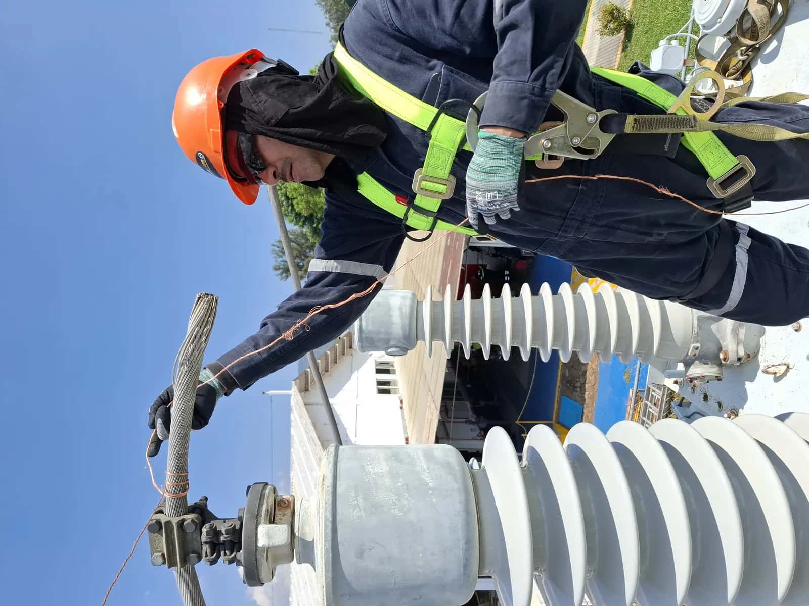 Técnico con arnés y casco inclinado sobre un aislador de alta tensión, sujetando el cable de conexión