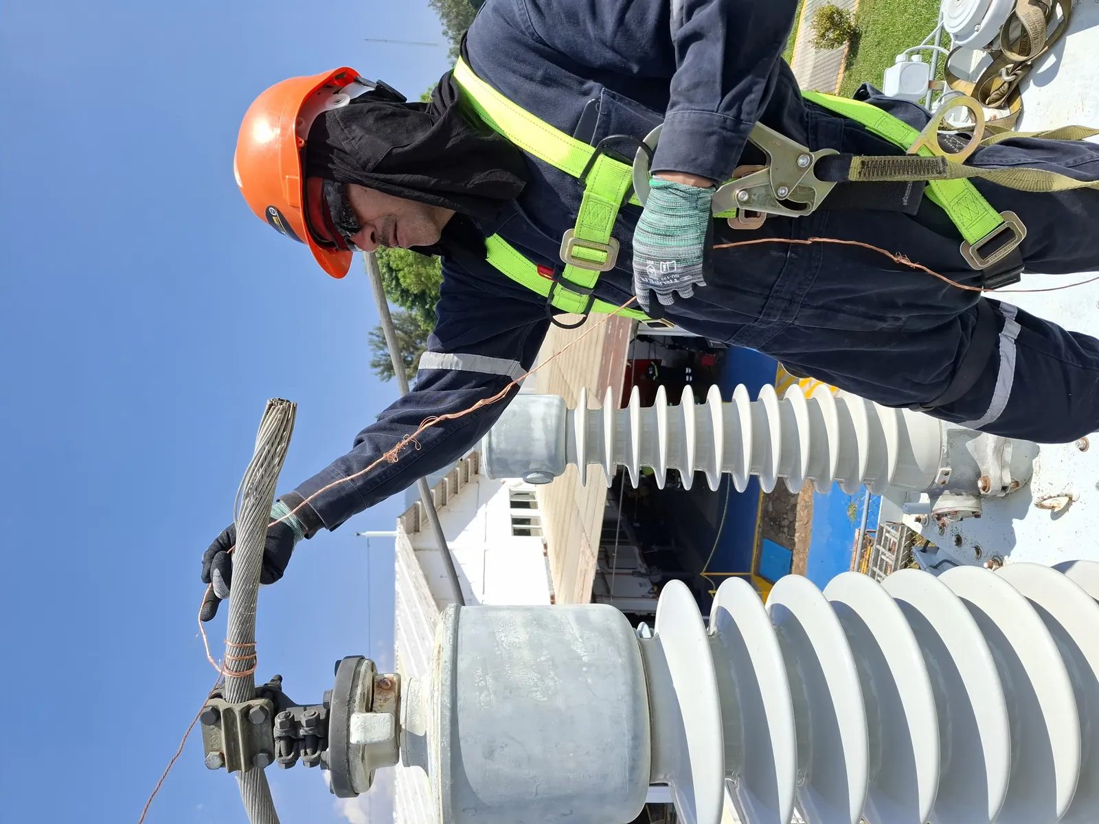Técnico con arnés y casco inclinado sobre un aislador de alta tensión, sujetando el cable de conexión
