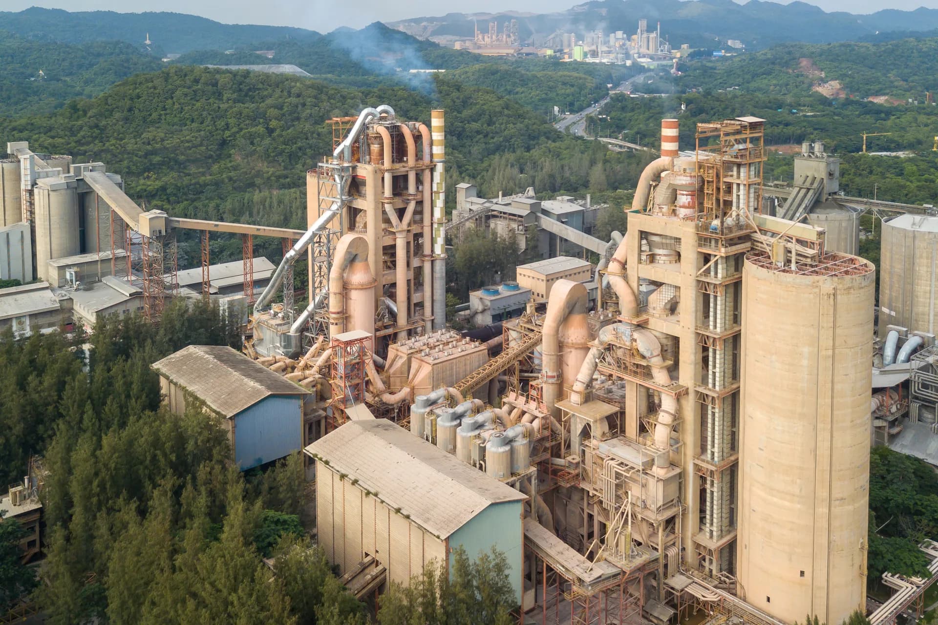 Planta cementera vista desde el aire, con torre de precalentamiento, silos y bandas transportadoras entre cerros boscosos.